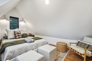 Un dormitorio moderno y bien iluminado, con techo inclinado, un ventanal grande, una cama con varias almohadas, una mesita de noche, una silla y una cesta tejida.