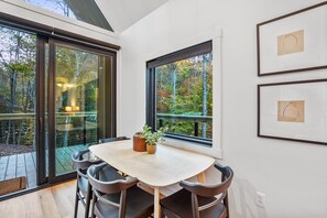 A dining area with a table and chairs, a window with a view of trees, and framed artwork on the wall. - Rocky Falls | Hocking Hills, Modern Cabin, Hot Tub (Rockbridge)