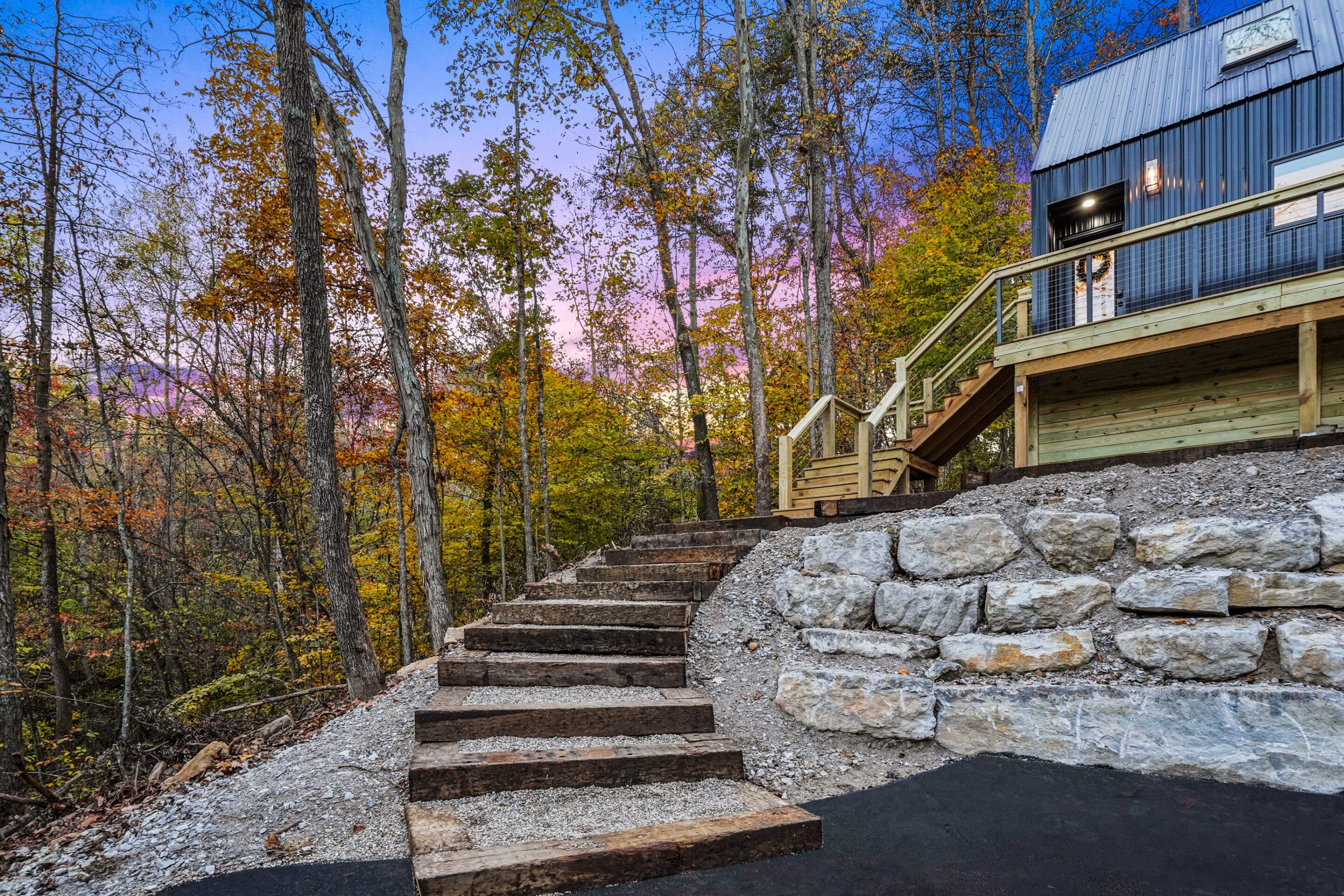 Un escalier menant à une terrasse en bois, bordée d’un mur de pierres et entourée d’arbres aux feuilles d’automne.