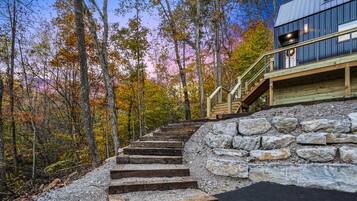 Un escalier menant à une terrasse en bois, bordée d’un mur de pierres et entourée d’arbres aux feuilles d’automne.