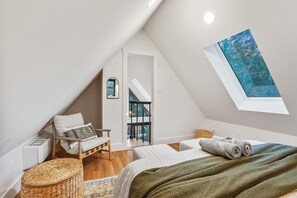 Une chambre moderne et bien éclairée, située sous les combles, avec un plafond en pente, une grande lucarne, un lit recouvert d'une couverture verte, une chaise en osier et une vue sur l'extérieur.