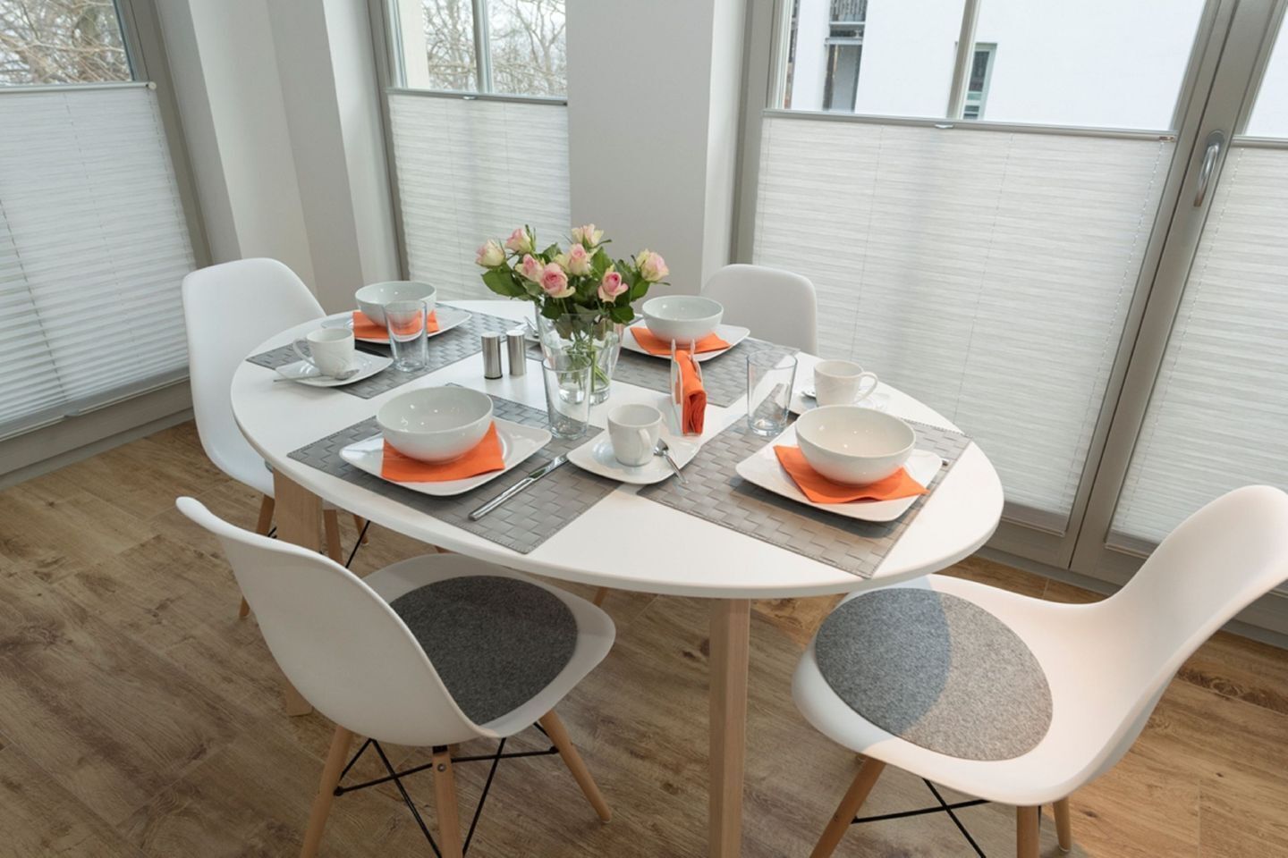 Un comedor moderno con una mesa redonda para cuatro personas, sillas blancas con cojines grises y un florero con flores.