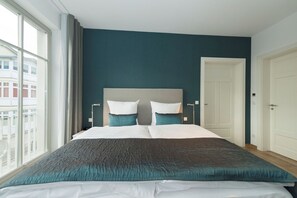 Habitación de hotel con una cama grande, dos mesitas de noche, cabecera y vistas de la ciudad a través de amplios ventanales.