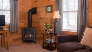 Un interior de cabaña acogedor con un estufa de leña, un televisor sobre una mesa de madera, un sofá y una mesita con libros y un jarrón de flores.