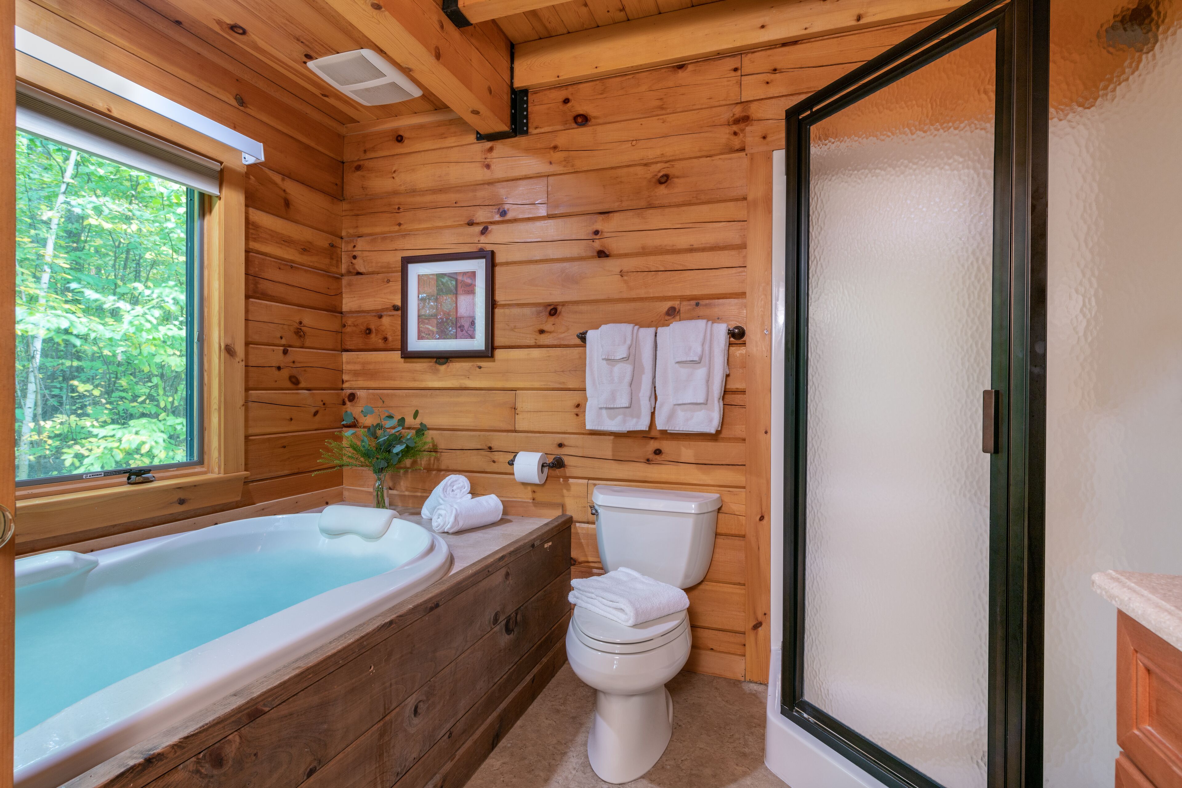 A wooden cabin bathroom with a bathtub, toilet, and a window with a view of trees.