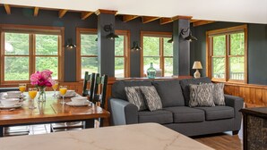 A dining area with a wooden table set for breakfast, a gray sofa with patterned cushions, and large windows with wooden frames.