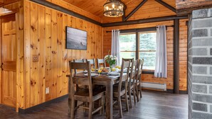 Une salle à manger aux murs lambrissés, avec une grande table dressée pour six personnes, un lustre et une fenêtre ornée de rideaux.