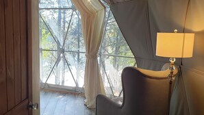 Ein Zeltraum mit Holztür, einem gepolsterten Sessel, einer Stehlampe und Blick auf Bäume durch ein großes Fenster.