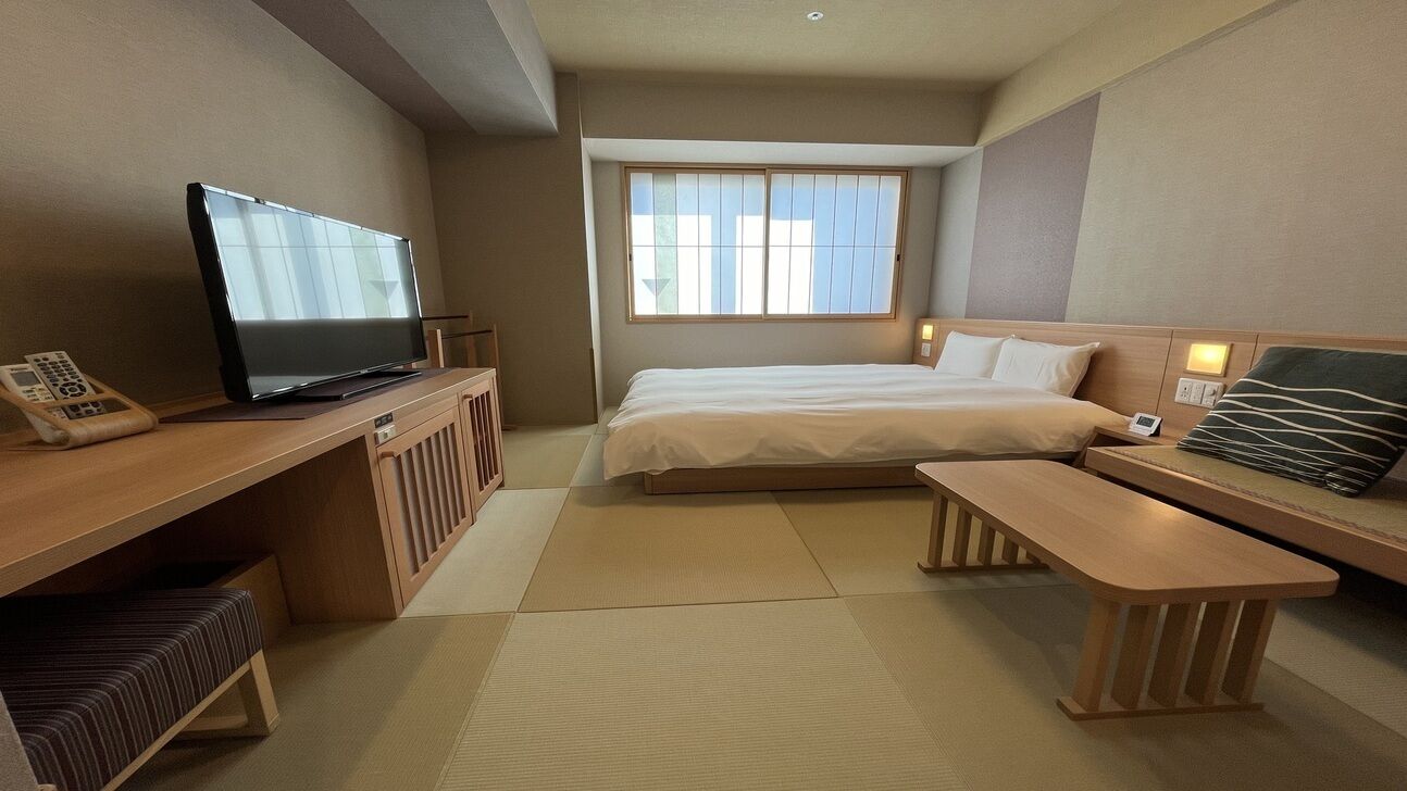 Een hotelkamer met een bed, een bureau met televisie, een nachtkastje en een raam met jaloezieën.