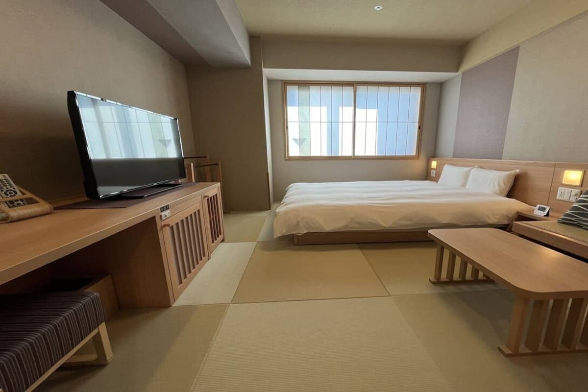 A hotel room with a bed, a desk with a television, a bedside table, and a window with blinds.