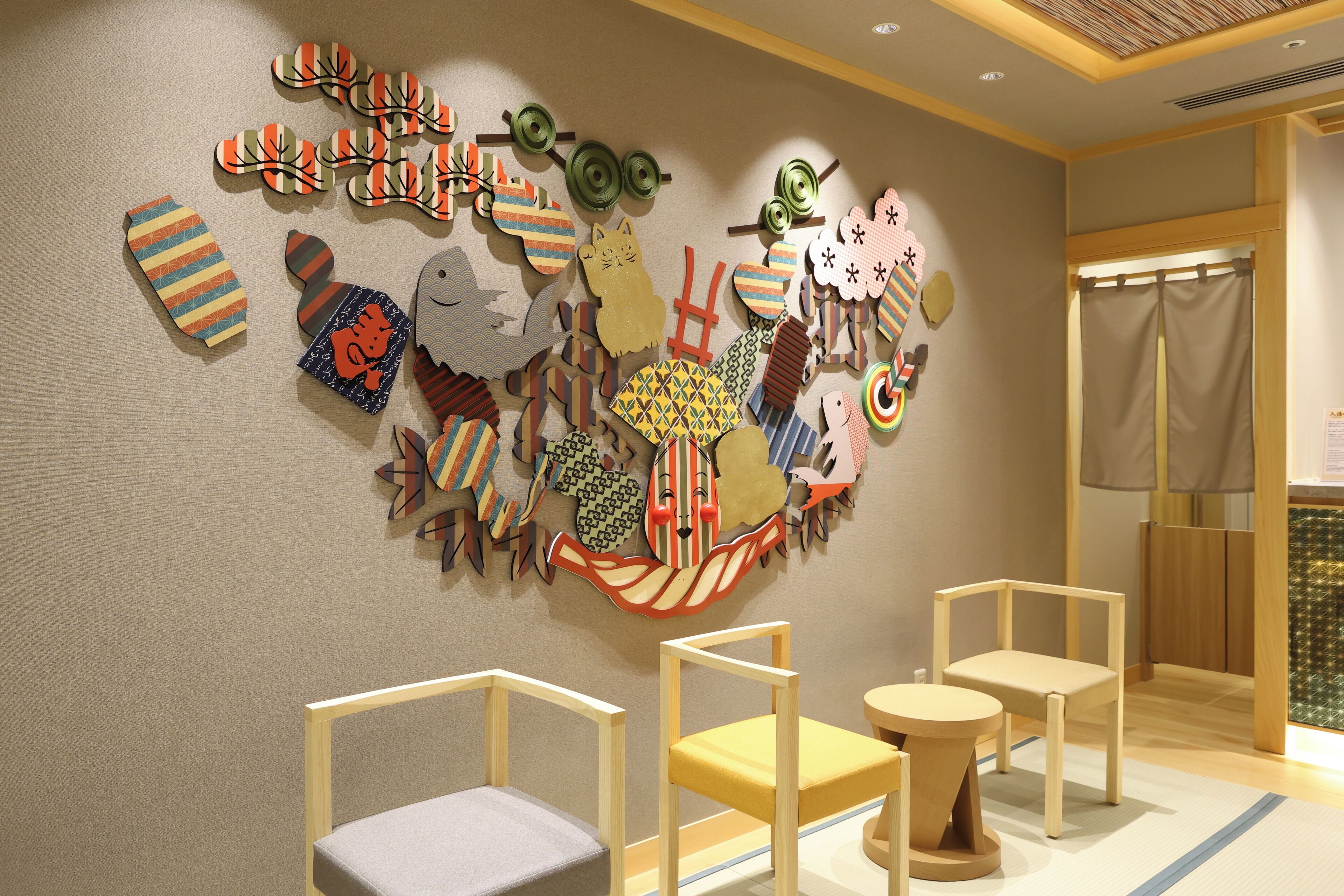 A room with a wall decorated with various colorful paper cutouts, including shapes and patterns. There are three chairs and a small table in the room.