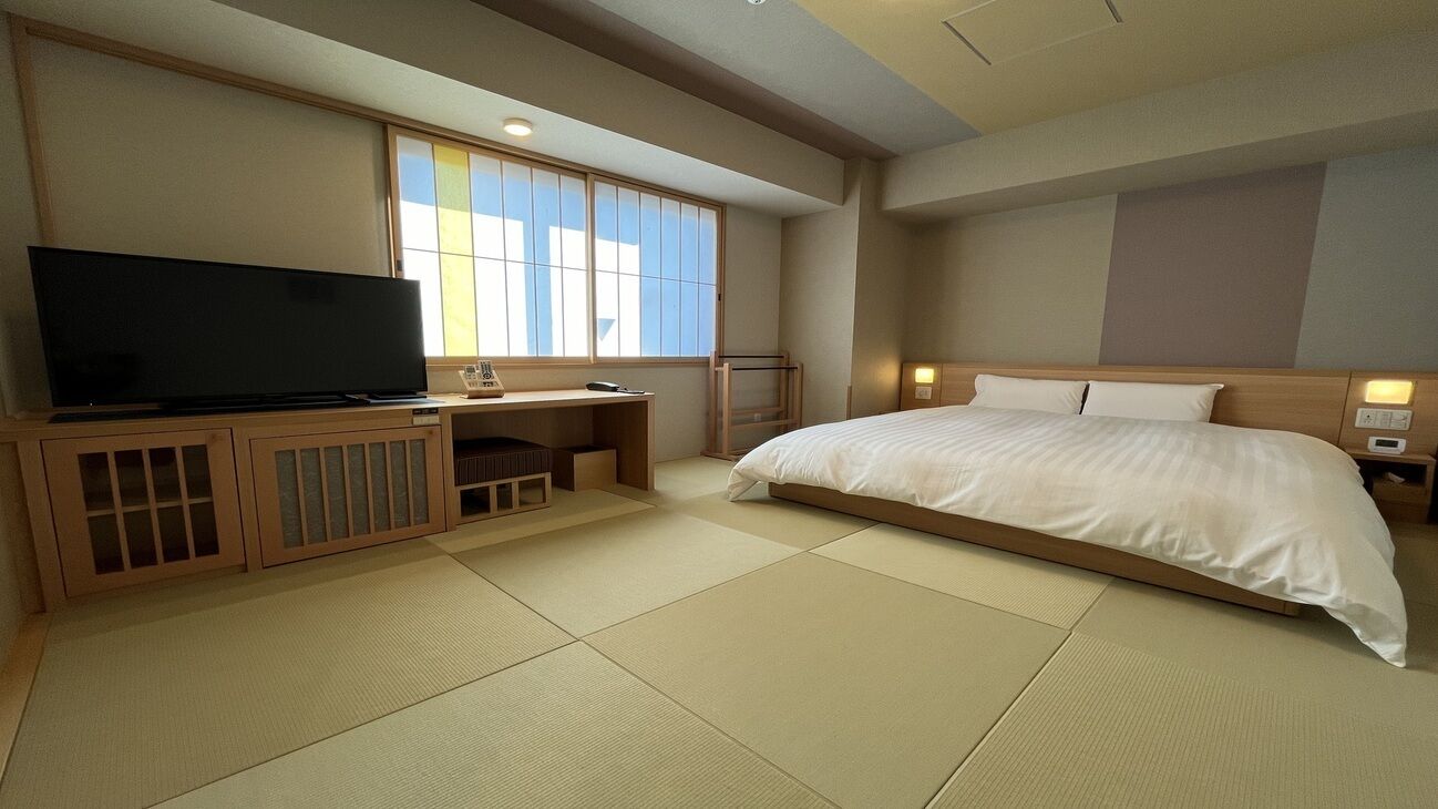 Een hotelkamer met tatami vloeren, een bed, een bureau met een televisie en een raam met uitzicht.