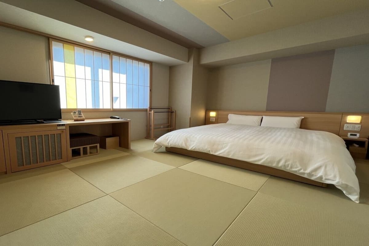 A hotel room with tatami flooring, a bed, a desk with a television, and a window with a view.