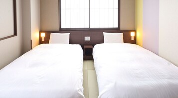 A hotel room with two single beds, a window with blinds, bedside lamps, and a power outlet on the wall.
