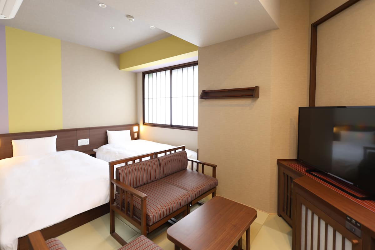 A hotel room with two beds, a sofa, a TV, and a window with blinds.