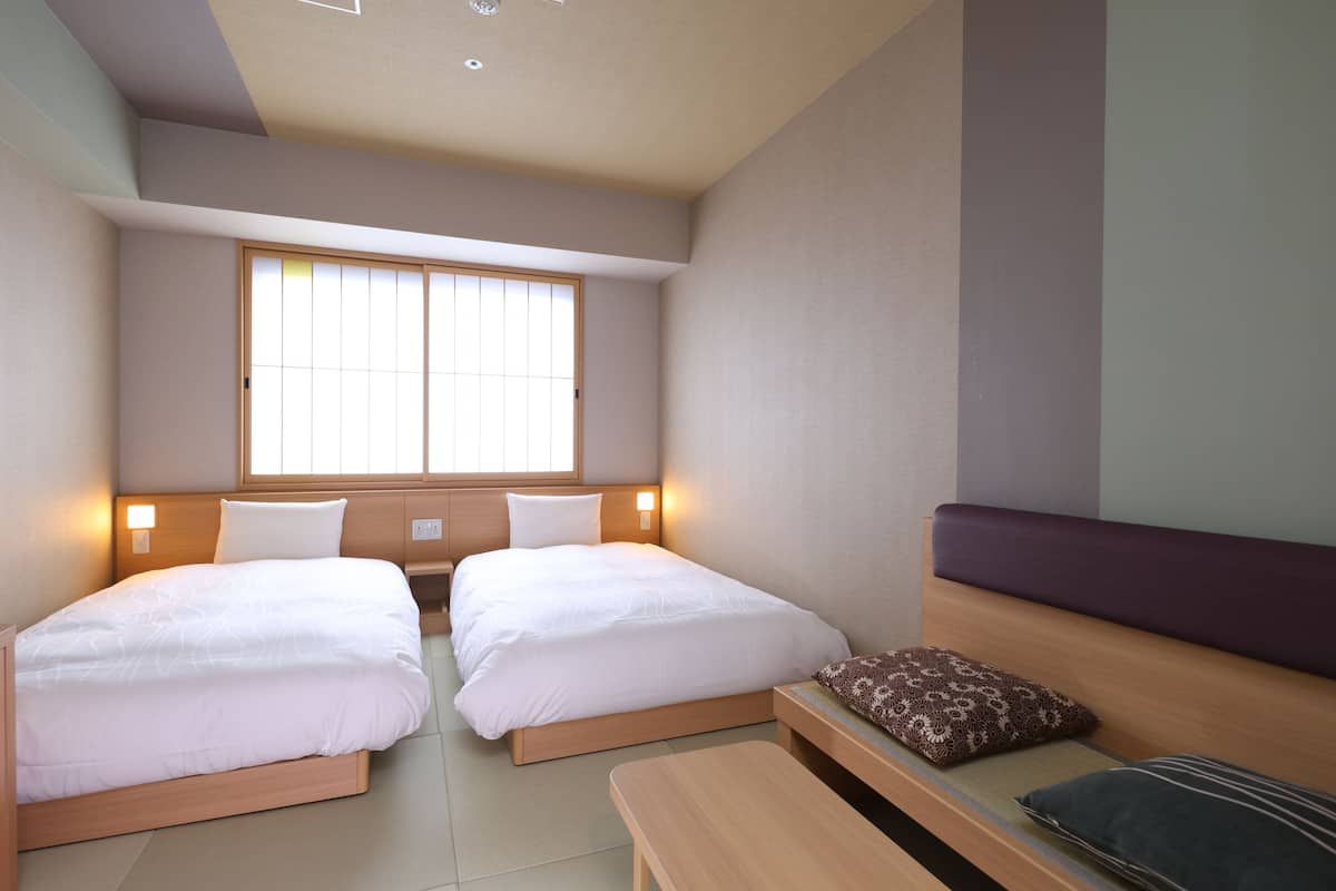 A hotel room with two beds, a window, a bedside table, and a bench.