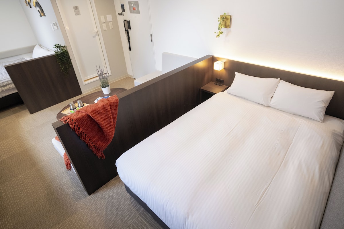 Quarto de hotel com uma cama grande, uma escrivaninha, uma cadeira, uma mesinha, uma mesa de cabeceira, uma luminária, uma planta e um ponto de luz fixado na parede.