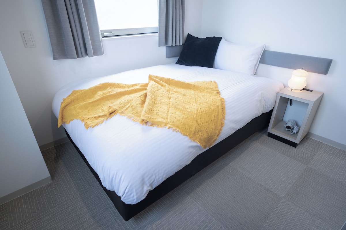 Cama bem arrumada com lençóis brancos e um cobertor amarelo, mesa de cabeceira com abajur e janela com cortinas.