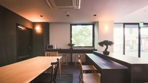 A modern cafe with a long wooden table, several chairs, a small bar area, and a large window with a view of the outside. - WAYFARER Matsu (Osaka)