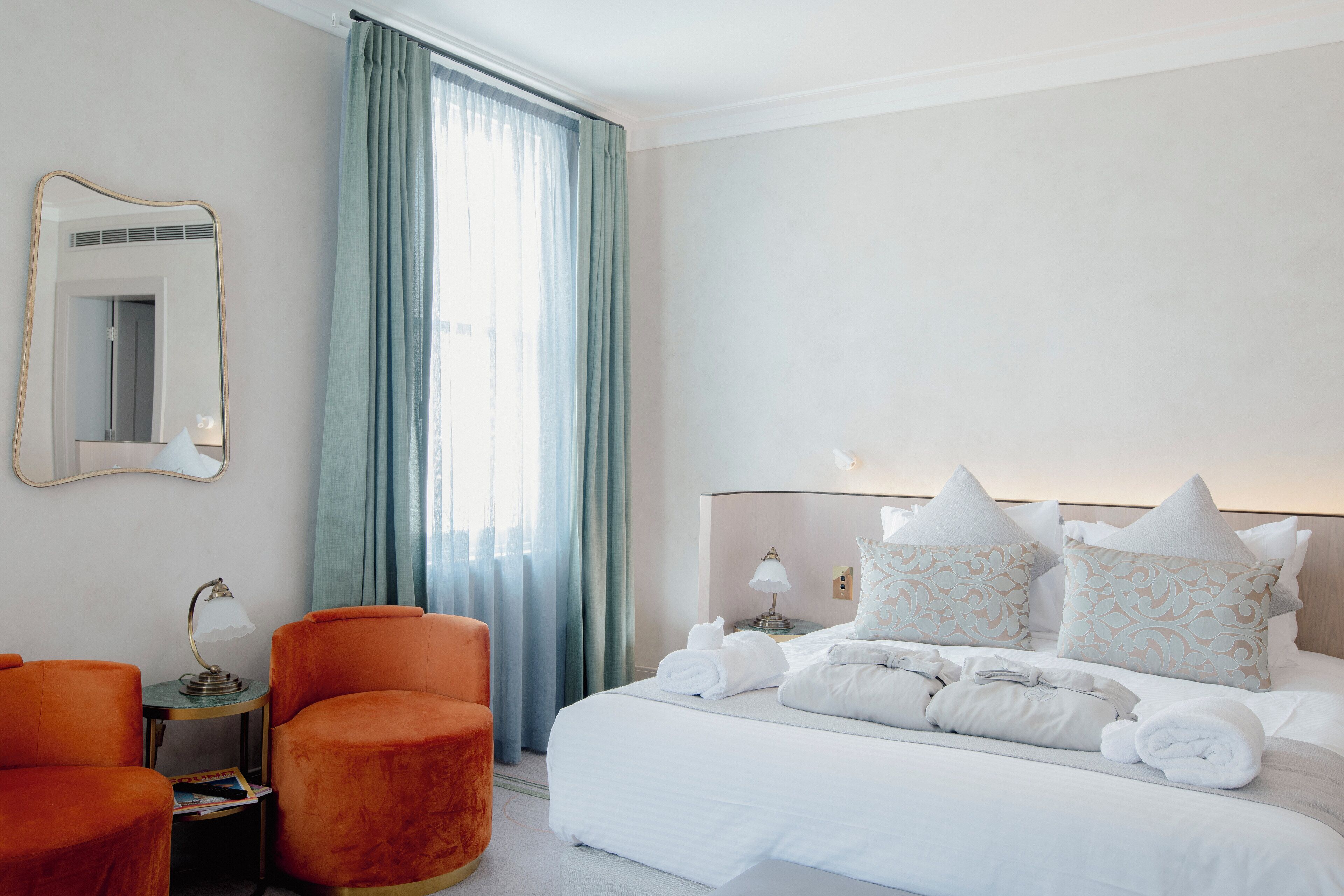 A hotel room with a neatly made bed, two armchairs, a small table with a lamp, and a large mirror on the wall.