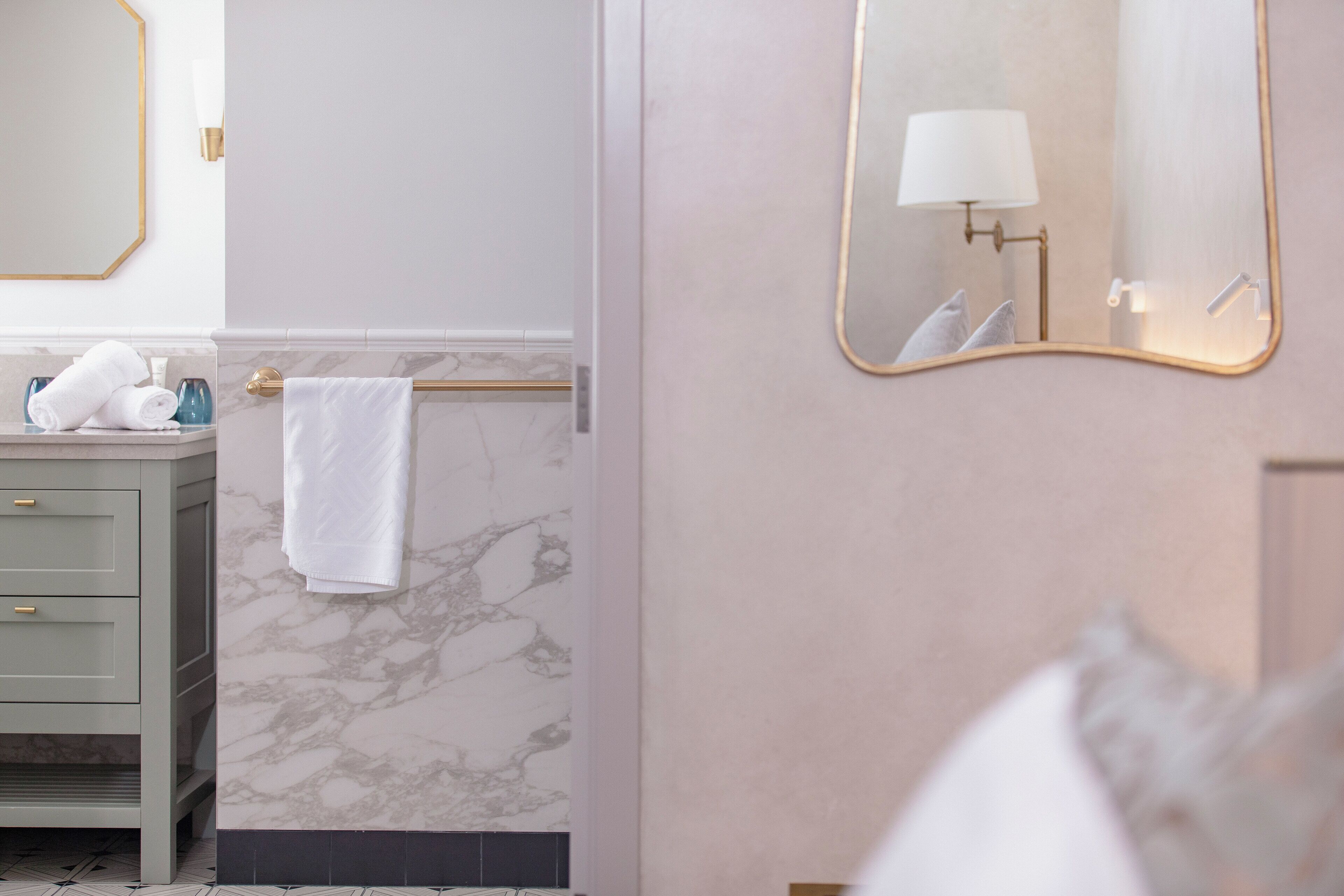 A modern bathroom with a marble wall, a gold-framed mirror, a white towel on a towel rack, and a green vanity with a towel dispenser.