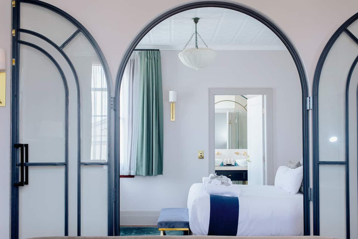 Una habitación de hotel con una cama, un banco, un espejo y vistas a través de un arco.