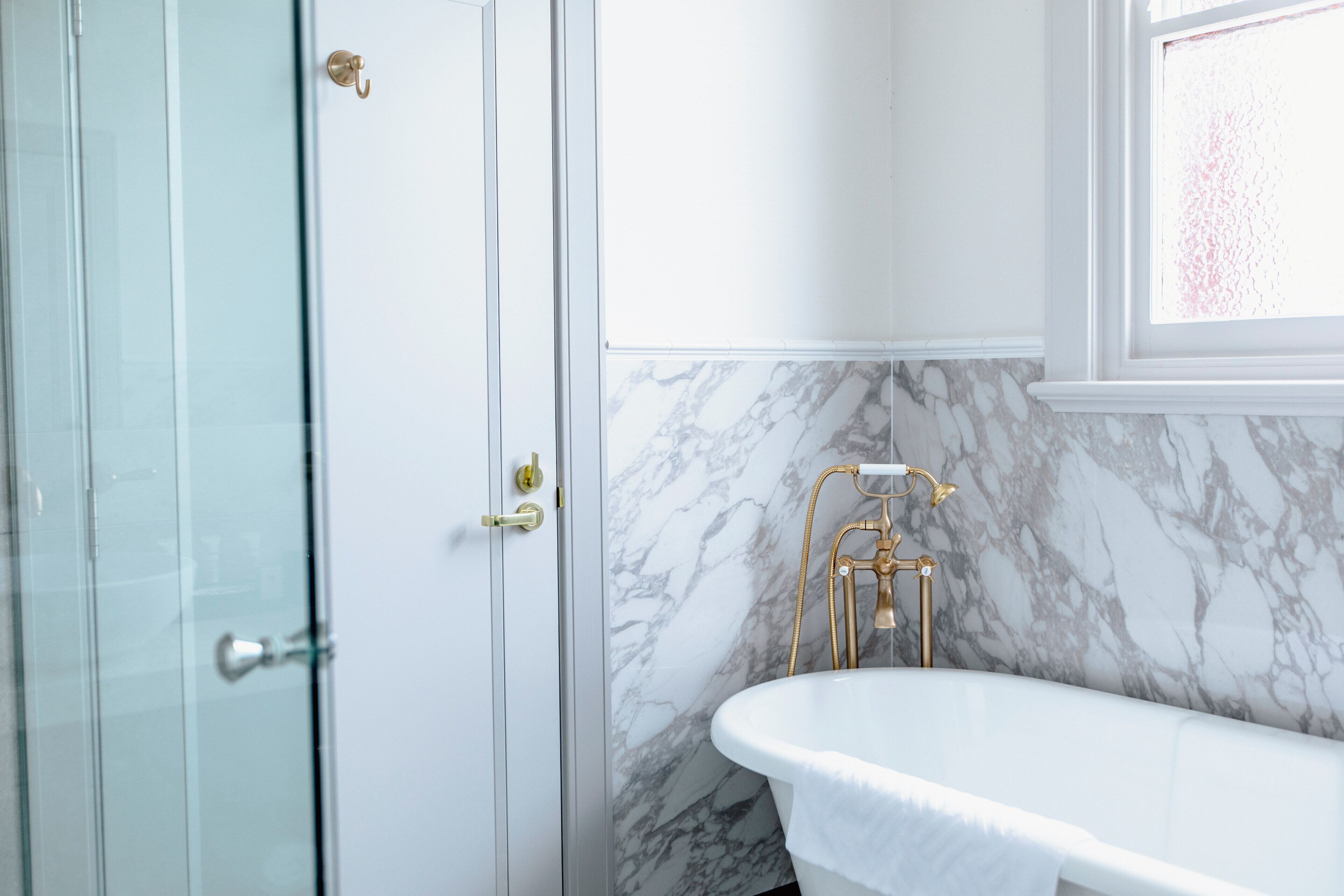 Un baño con cabina de ducha de vidrio, una bañera blanca con detalles dorados y paredes de mármol.