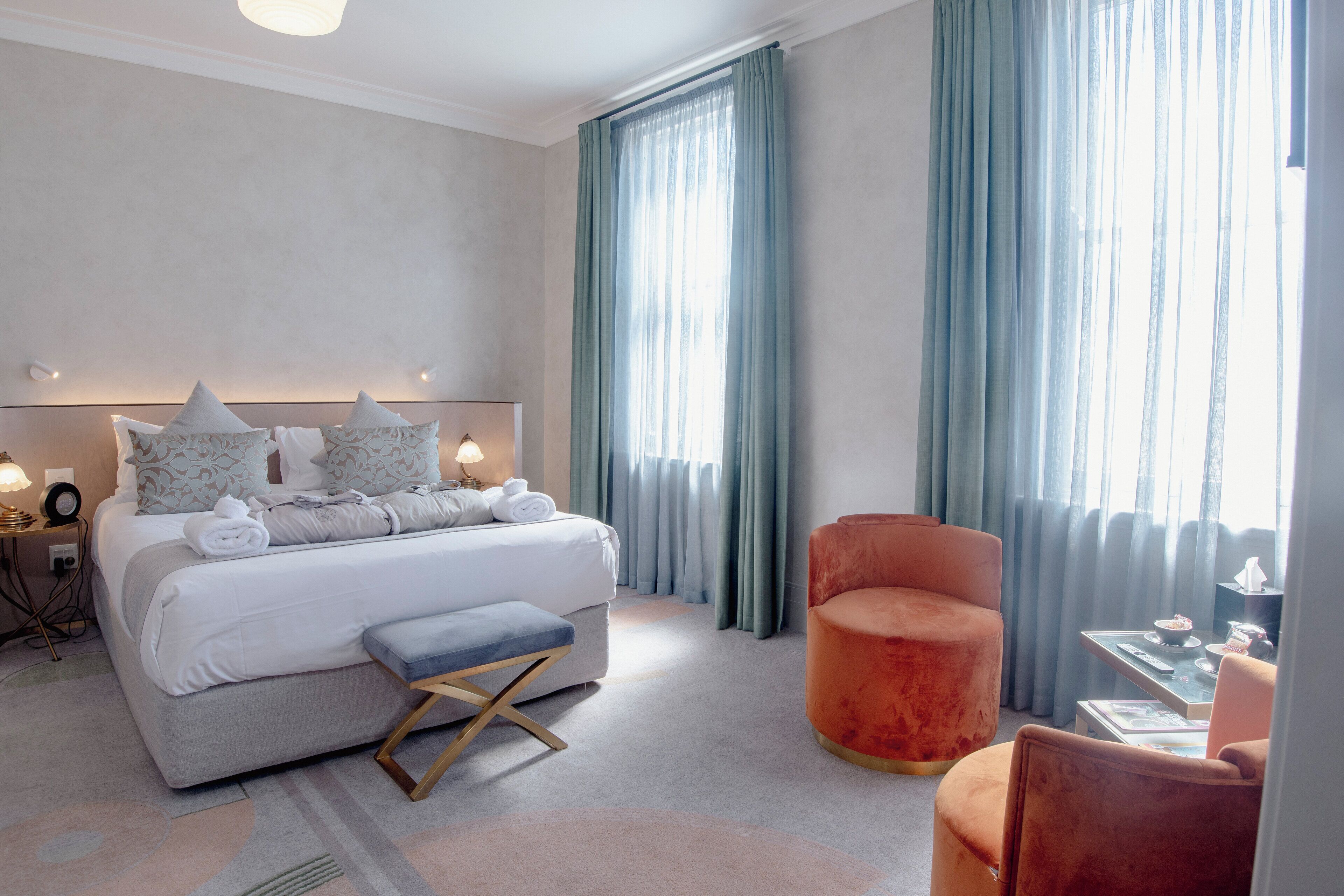 A modern hotel room with a large bed, a bench, a red armchair, and a small table with a bowl and a bottle.