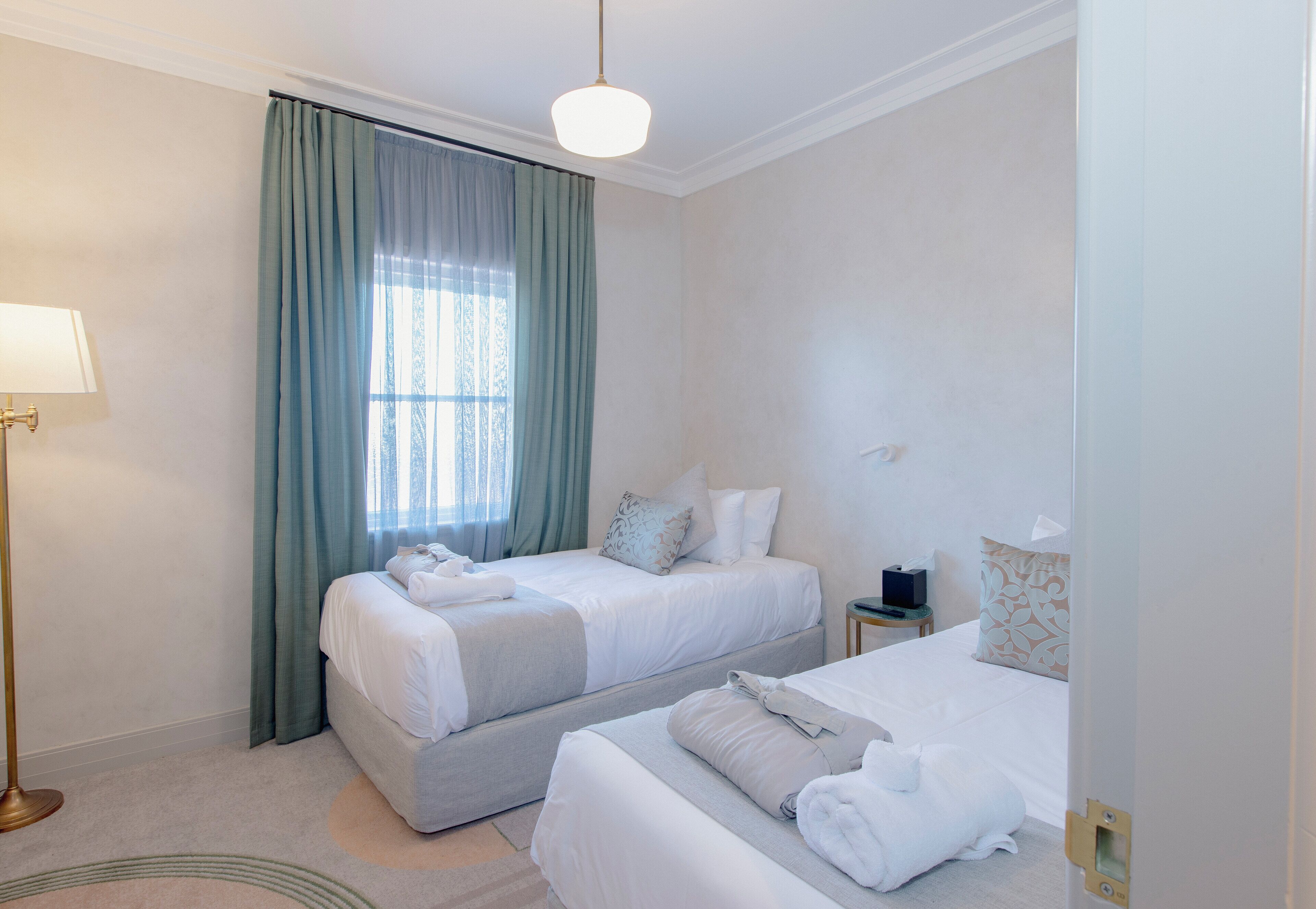 Quarto de hotel com duas camas, uma mesinha de cabeceira, um abajur e uma janela com cortinas.