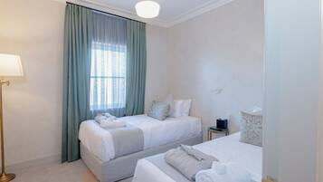Quarto de hotel com duas camas, uma mesinha de cabeceira, um abajur e uma janela com cortinas.