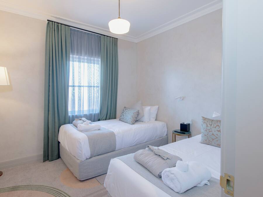 Quarto de hotel com duas camas, uma mesinha de cabeceira, um abajur e uma janela com cortinas.
