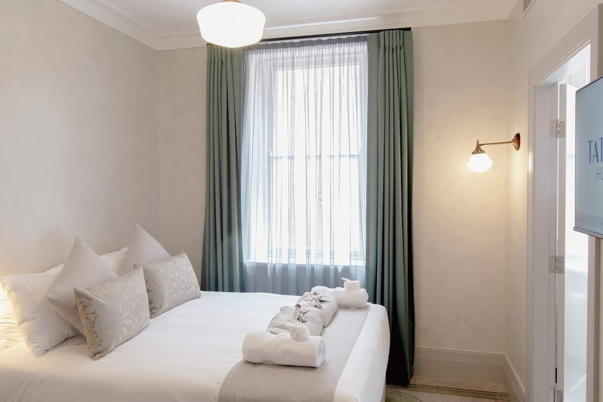 Una cama impecablemente arreglada con sábanas blancas y almohadas decorativas, un ventanal con cortinas traslúcidas y una lámpara fijada a la pared.
