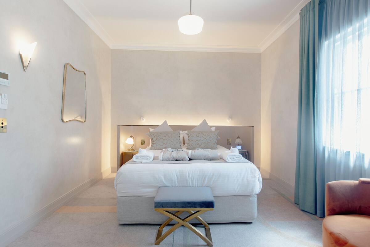 Un dormitorio ordenado con una cama grande, lámparas de noche, una mesita de noche, un banco y una silla.