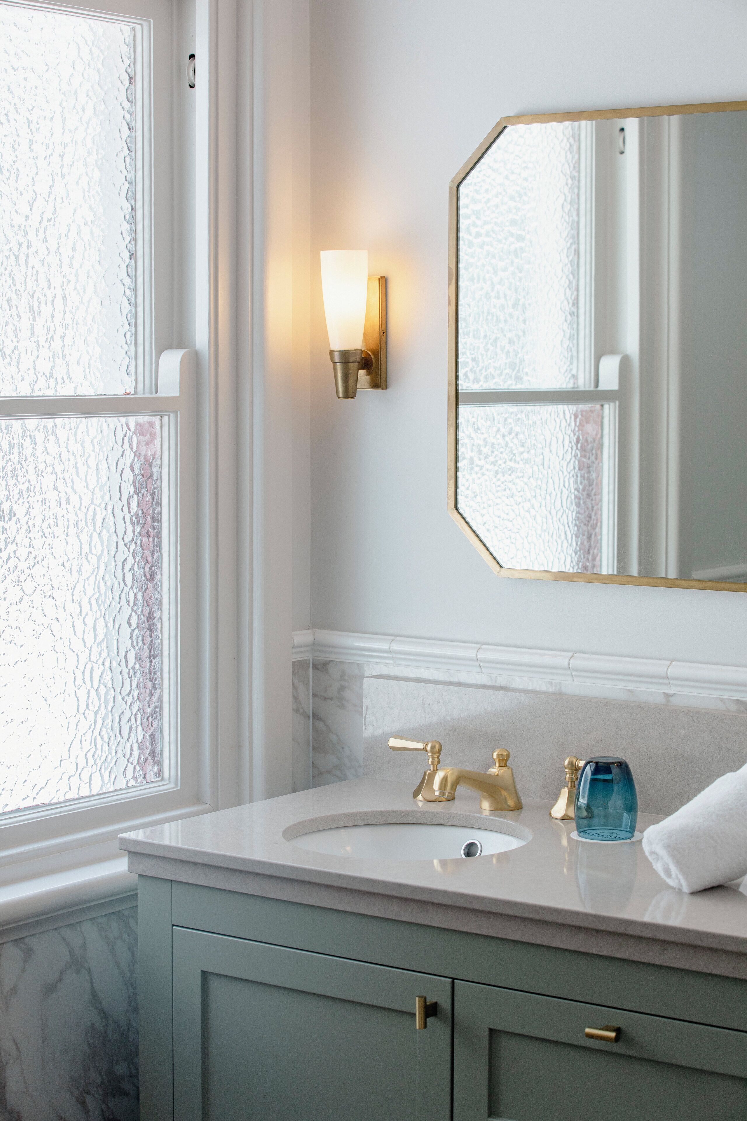 A bathroom with a marble sink, gold fixtures, a wall-mounted mirror, and a frosted glass window.
