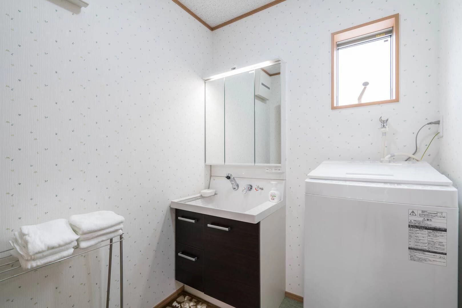 Ein kleines Badezimmer mit einem weißen Waschbecken, einem dunklen Schrank und einem Spiegel darüber. Eine Handtuchstange mit Handtüchern und ein kleines Fenster mit Holzrahmen sind ebenfalls vorhanden.