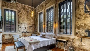 Une chambre avec un grand lit, un bureau et une chaise. Il y a trois fenêtres équipées de volets.