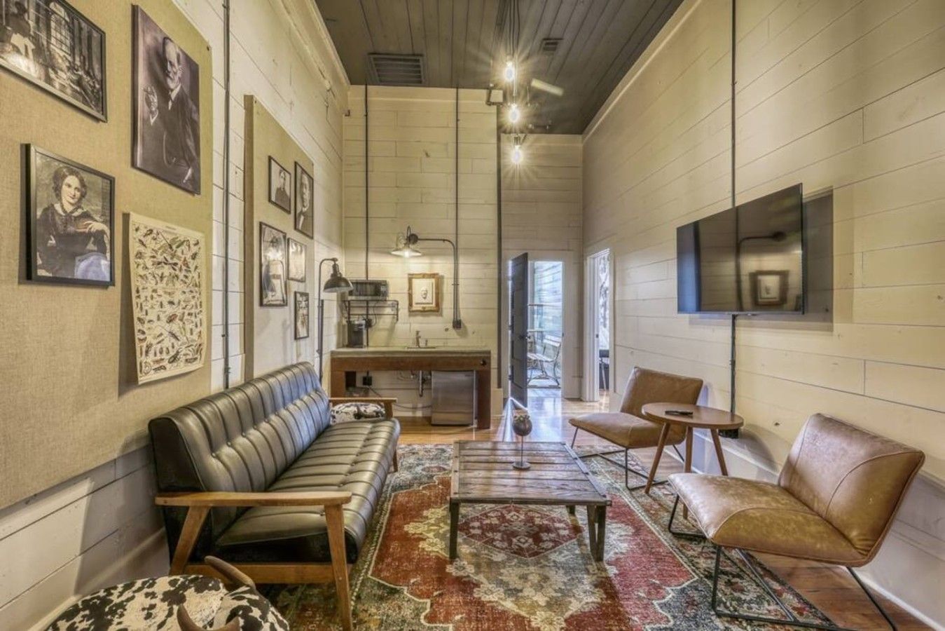 A spacious living room with a leather sofa, a coffee table, and a rug. There are framed pictures on the walls and a television mounted on the wall.