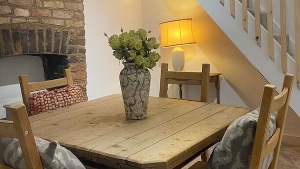 Un comedor con una mesa y sillas de madera, un jarrón con plantas verdes, una pared de ladrillo y una escalera con barandilla.