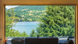 A room with a large window overlooking a lake and hills. There is a sofa with a blanket and a hat on it. - Veranda 4M  property (Colibita)