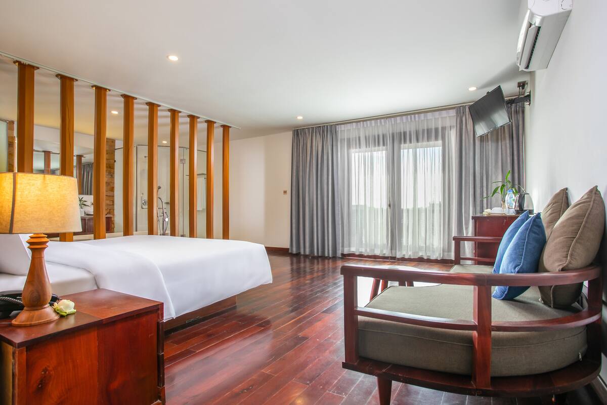 Una habitación de hotel con una cama, una silla de madera, un escritorio con lámpara, un ventanal con cortinas y un equipo de aire acondicionado.