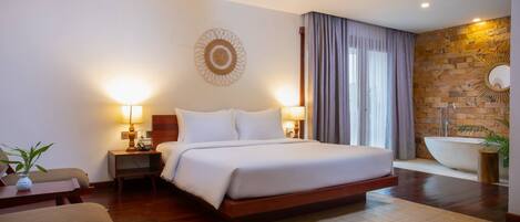 A hotel room with a large bed, bedside lamps, a small table with a plant, a wooden floor, a large mirror, and a bathtub.