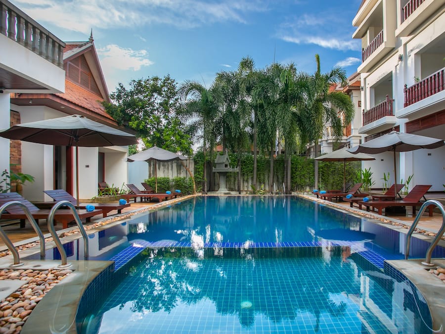 Piscina con sillones de descanso y sombrillas, rodeada de edificios con balcones.