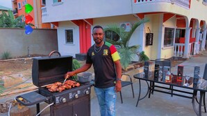 Ein Mann grillt draußen, ein Grill, ein Tisch mit Flaschen und ein Gebäude im Hintergrund.