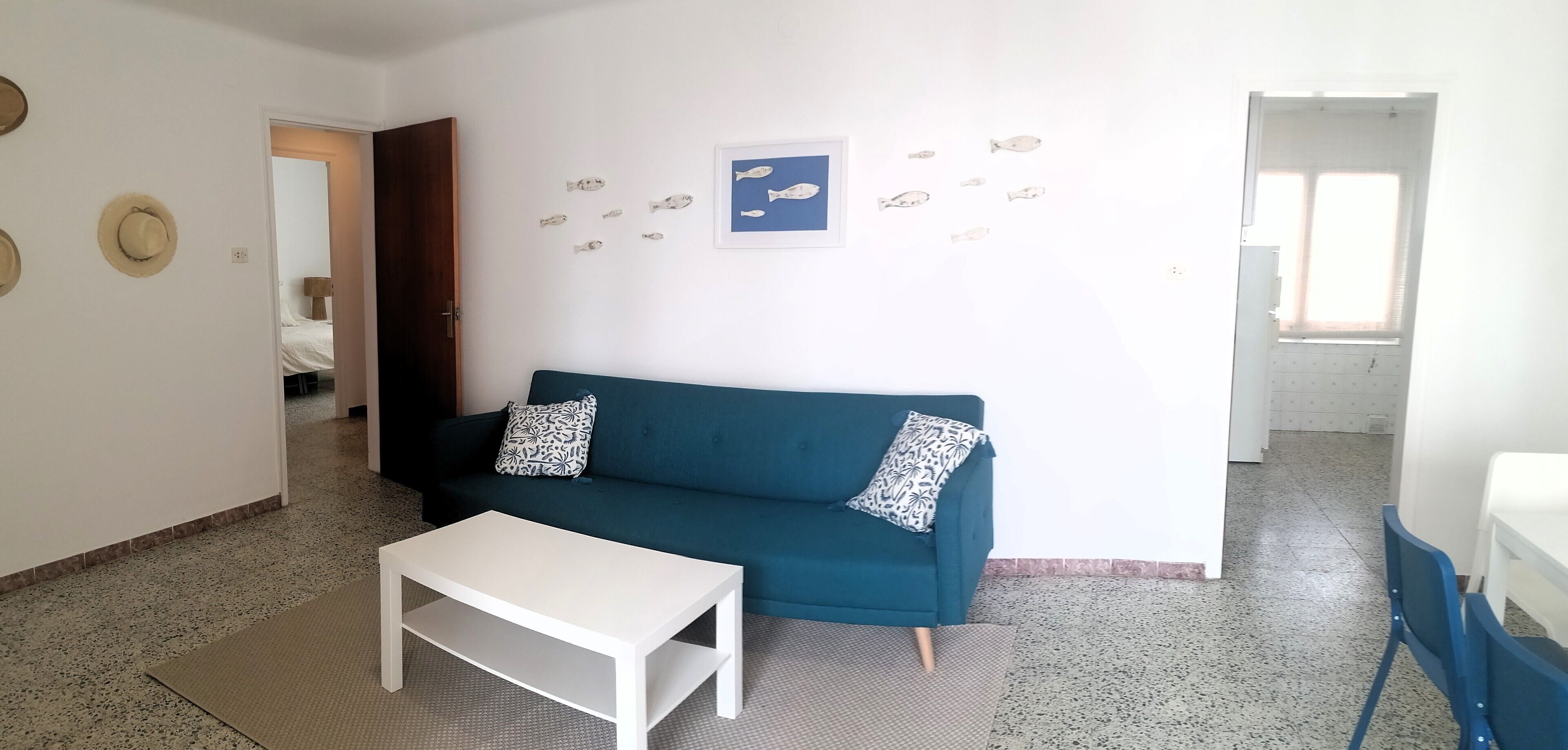 Een woonkamer met een blauwe bank, een witte salontafel en gekleurde kussens. Er zijn decoraties aan de muur van vissen en een ingelijste foto.