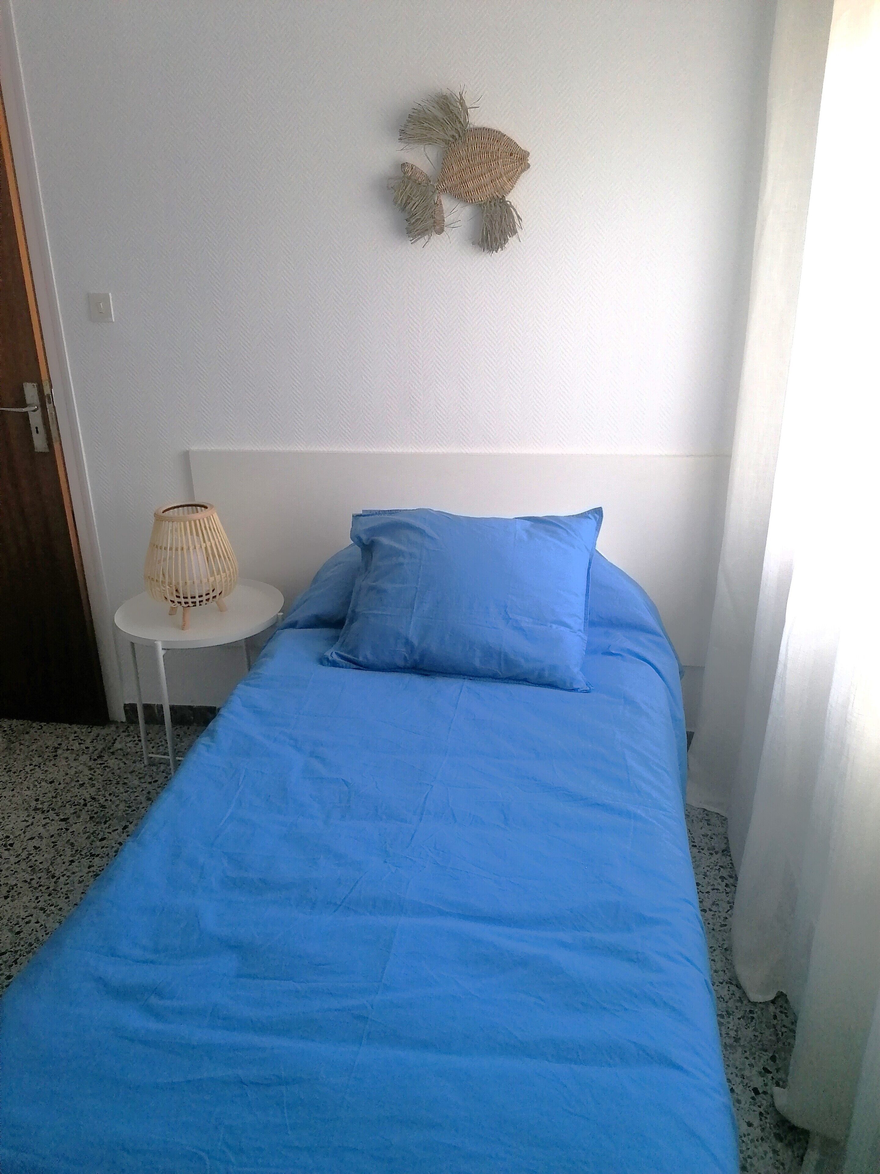 A single bed with a blue bedspread and pillows, a small round table with a woven lamp, and a wall decoration.