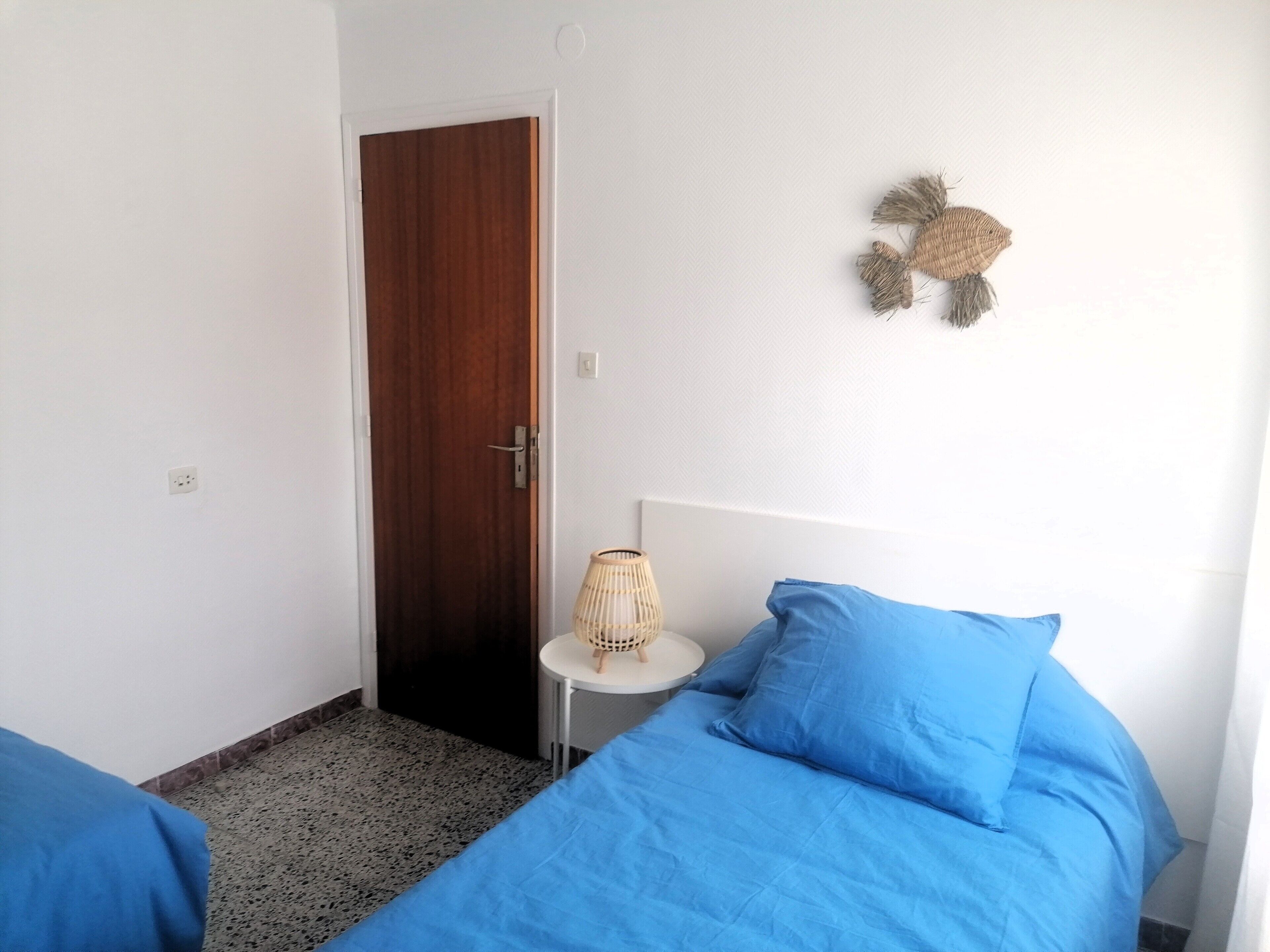 A bedroom with a wooden door, a small round table with a woven lamp, and two blue beds.