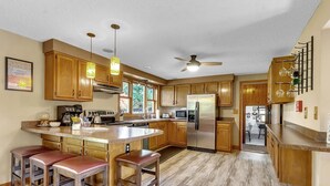 Una cocina con armarios de madera, una isla central con taburetes, un ventilador de techo y vistas a un comedor.