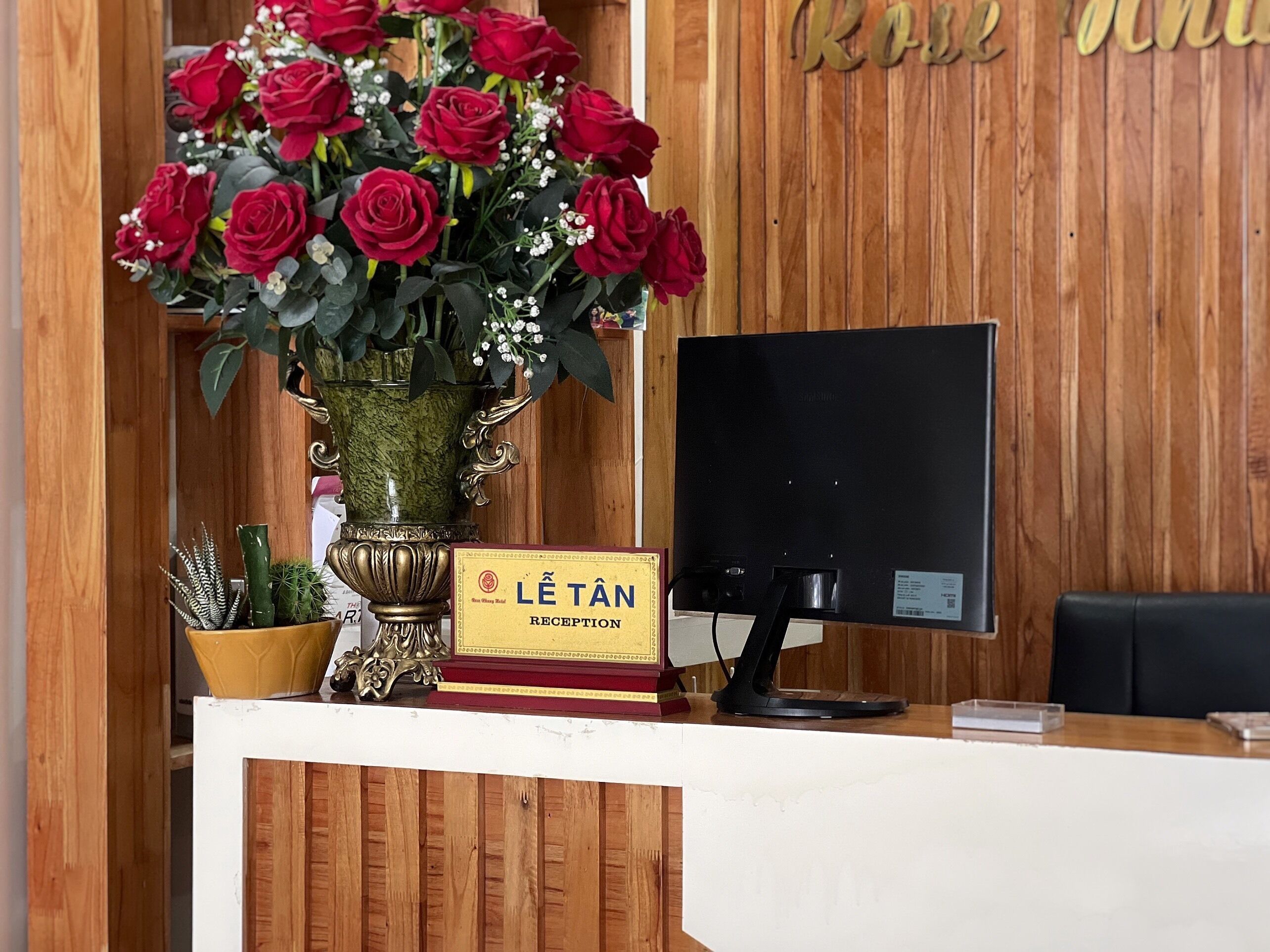 Ein Empfangsschalter mit einer großen Vase roter Rosen, einem Schild mit der Aufschrift 'Lê Tân Reception' und einem Computermonitor.