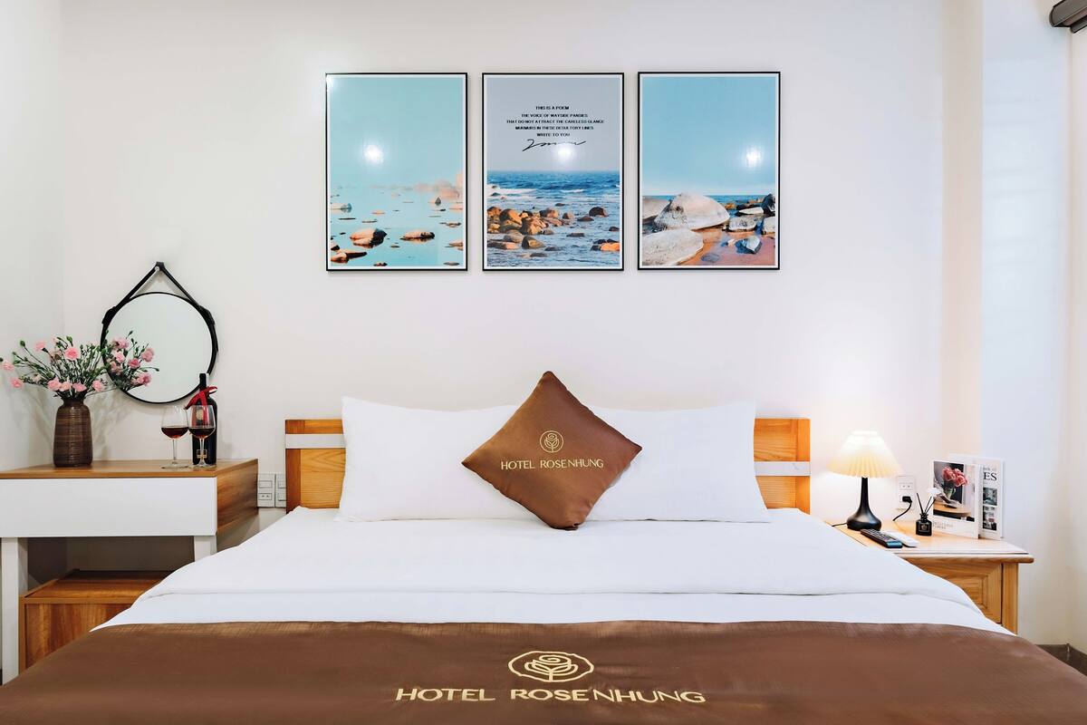 Une chambre d’hôtel avec un lit, une table de chevet, une lampe, un vase de fleurs et un poème encadré accroché au mur.