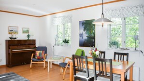 A living room with a piano, a dining table with chairs, a sofa, and a large window with curtains. - Cozy home in ålem with kitchen (Ålem)